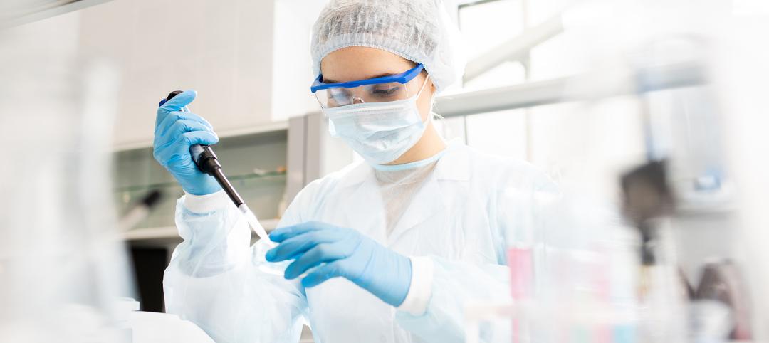 Serious concentrated female microbiologist in sterile clothing and safety goggles sitting at table and dropping reagent in petri dish while doing research in laboratory